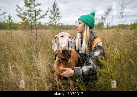 Ruhige Besitzerin umarmen den geliebten Vizsla-Hund, der nebeneinander in trockenem Seggengras sitzt, sich nach einem Spaziergang auf Torfmoor ausruht, in die Ferne blickt und die herbstliche Stille der nördlichen Natur genießt. || Modell freigegeben Stockfoto