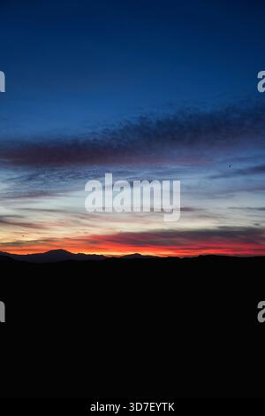 Lebendiger Sonnenuntergangshimmel mit Wolken, die in Rot-, Orange- und Blautönen über dunklen Bergsilhouetten leuchten, schaffen eine dramatische, ruhige Landschaft. Stockfoto