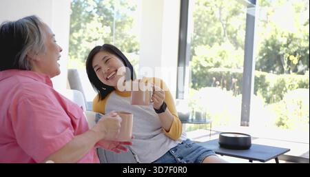 Plaudernde Frauen in rosa Hemd und Jeansshorts auf dem Sofa in der Nähe von Glastüren, die Tassen halten Stockfoto