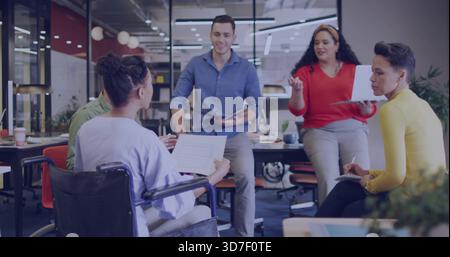 Vier Teammitglieder im Open Office prüfen Zeitungen, Rollstuhl, Laptop, Red Top Stockfoto