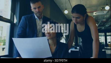 Sitzende Frau in dunklem Blazer mit gedrucktem Dokument im modernen Büro mit Teambeuge studieren Stockfoto