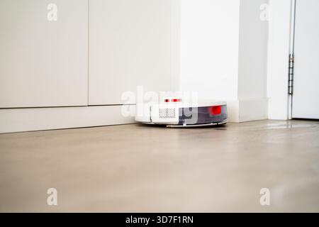 Roboter-Bodensauger, der entlang der Wandplatte arbeitet, während Kanten in einer hellen, minimalistischen Wohnumgebung gereinigt werden Stockfoto