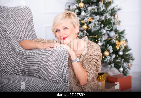 Frau mit kurzen blonden Haaren, in einem gemütlichen Pullover gekleidet, posiert neben einem mit Ornamenten geschmückten Weihnachtsbaum und schafft so eine fröhliche Urlaubsatmosphäre Stockfoto