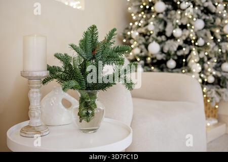 Festliche Kerzen- und Tannenzweige in Glasvase mit verschwommenen Weihnachtsbaumlichtern in gemütlichem Wohnraum, ideal für saisonales Design und Marketing. Stockfoto