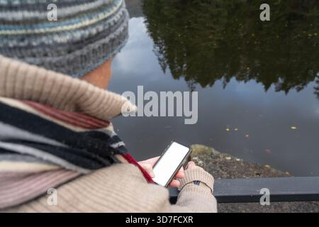 Die Person in gemütlicher Kleidung ist mit dem Smartphone in der Nähe des ruhigen Wassers beschäftigt, mit Bäumen und Reflexionen, die eine friedliche Atmosphäre für Entspannung und Entspannung schaffen Stockfoto