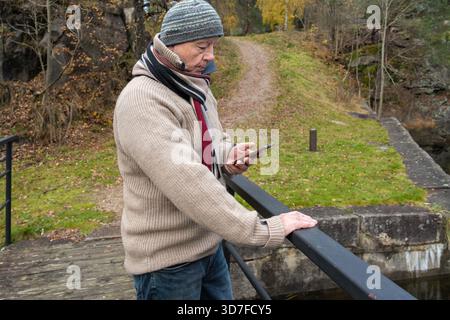 Ein älterer Mann in warmer Kleidung, der an einem Geländer steht, in einer ruhigen Umgebung im Freien mit herbstlichen Farben und einem natürlichen Klaps mit dem Smartphone beschäftigt ist Stockfoto