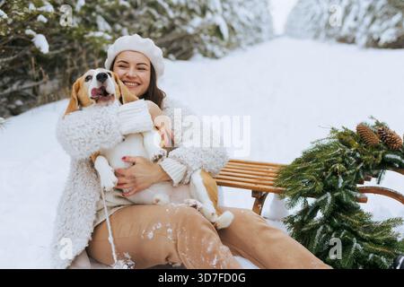 Fröhliche junge Frau kuschelt ihren Beagle-Hund auf einem Schlitten in einem verschneiten Wald, genießt Winterferien und gemütliche Outdoor-Zeit zusammen Stockfoto