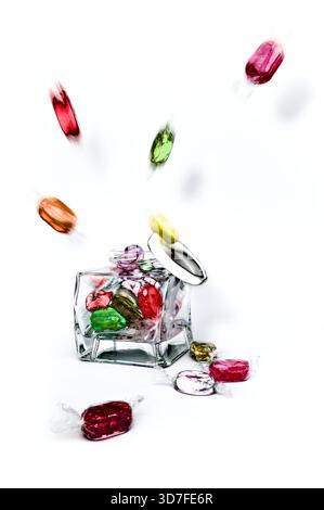 Studio-Aufnahme von bunten gekochten Süßigkeiten, die aus einem Glasgefäß aufplatzen, wobei der Deckel vor weißem Hintergrund mit künstlerischem Effekt abfällt. Stockfoto