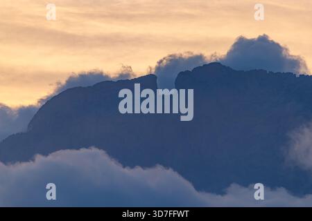 Eine Nahaufnahme der dunklen, schroffen Silhouette des Gipfels von Puig Campana, die „El Portell“ (die Spaltung) hervorhebt, eingerahmt von weichen, tief hängenden Wolken und Stockfoto