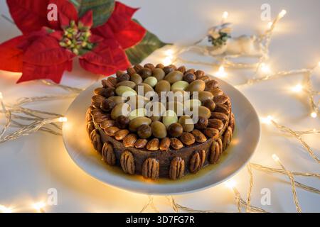 Traditioneller Weihnachts-Fruchtkuchen, dekoriert mit Pekannüssen, schokoladenüberzogenen Paranüssen und Aprikosen-Glasur auf einem weißen Teller mit festlichen Lichterketten. Stockfoto