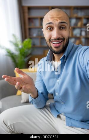 Junger kahlköpfiger Mann lächelt und gestickt während eines freundlichen Vlog- oder Videoanrufs von seiner modernen Wohnzimmercouch aus, lässiges blaues Hemd, selbstbewusst und ansprechend vor der Kamera Stockfoto