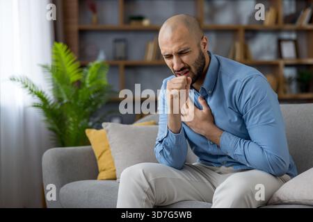 Junger Mann, der zu Hause auf einer Couch sitzt, seine Brust anklammert und Husten, zeigt Atemwegserkrankungen oder allergische Reaktionen mit Unwohlsein und Müdigkeit in einem Wohnzimmerbereich Stockfoto