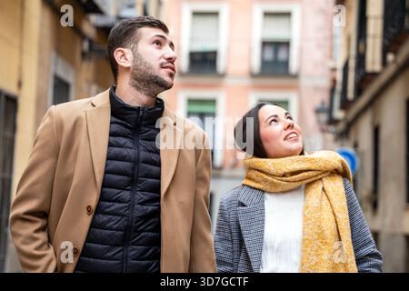 Ein junges Paar schlendert durch eine lebhafte Straße in Madrid und bestaunt die atemberaubende Architektur oben. Es ist ein kühler Herbsttag und warme Farben Surro Stockfoto