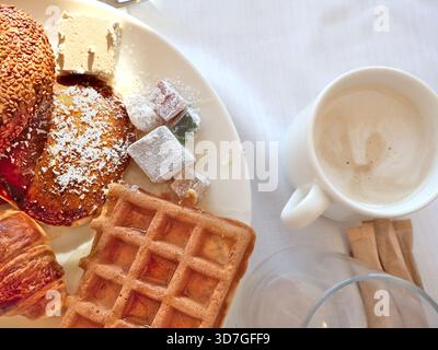 Eine herzhafte Morgenszene mit Waffeln, Croissants, Sesambrötchen und verschiedenen Konfekt sowie einer cremigen Tasse Kaffee auf einem hellen weißen Tisch. Stockfoto
