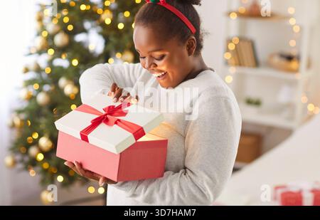 Frohe Weihnachten und frohe Neujahrsfeiertage neugierige afroamerikanerin, die Geschenkbox öffnet Stockfoto