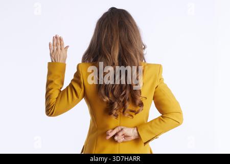 Eine professionelle Frau in einem gelben Blazer macht mit einer Hand eine Geste der Aufrichtigkeit, während sie diskret die Finger hinter ihrem Rücken drückt und einen fals signalisiert Stockfoto