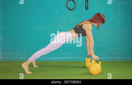 Frau, die eine vordere Planke mit zwei gelben Kettlebells vorführt, wobei ihre Beine vollständig ausgestreckt bleiben. Stockfoto