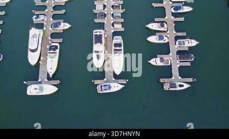 Bei einer Drohnenaufnahme über der Decke werden viele Freizeitboote und Yachten sichtbar, die in geordneten Reihen an einem ruhigen Yachthafen liegen, umgeben von ruhigem, tiefgrünem Wasser. Stockfoto