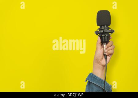 Nahhand, die ein schwarzes Mikrofon vor einem leuchtend gelben Hintergrund hält, symbolisiert Podcasting, Audioaufnahme, Übertragung, Medienproduktion, Stockfoto