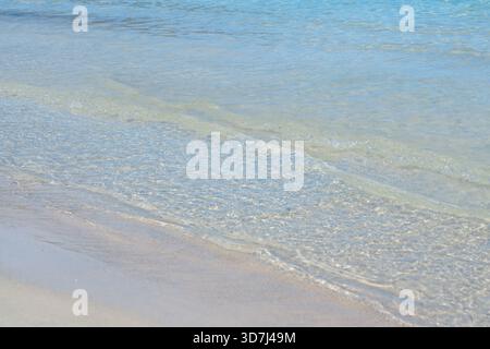 Sanfte Wellen ziehen über eine flache Sandküste und enthüllen die Klarheit des türkisfarbenen Meerwassers. Die weiche Textur und das natürliche Licht sorgen für eine ruhige Atmosphäre Stockfoto