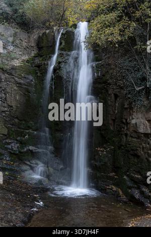 Dieses fesselnde Bild zeigt einen ruhigen Wasserfall, der durch felsige Klippen stürzt, umgeben von üppigem Grün und Herbstlaub, was eine friedliche Atmosphäre schafft Stockfoto