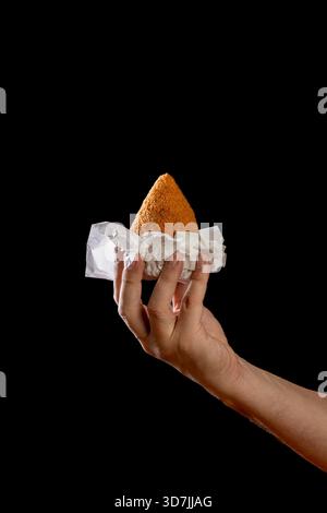 Hand mit einem traditionellen sizilianischen Arancino. Nahaufnahme der berühmten frittierten Reiskugel, ein typisches Straßenessen Siziliens, isoliert auf einem dunklen stimmungsvollen B Stockfoto