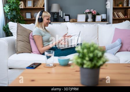 Lächelnde junge Frau mit Kopfhörern, die auf dem Sofa sitzen, während sie mit dem Laptop Musik und entspannter Bildschirmzeit beim Umschalten zwischen Online-Hengst genießt Stockfoto