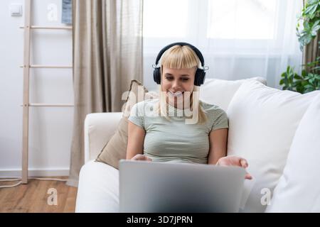 Lächelnde junge Frau mit Kopfhörern, die auf dem Sofa sitzen, während sie mit dem Laptop Musik und entspannter Bildschirmzeit beim Umschalten zwischen Online-Hengst genießt Stockfoto