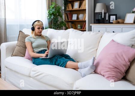 Lächelnde junge Frau mit Kopfhörern, die auf dem Sofa sitzen, während sie mit dem Laptop Musik und entspannter Bildschirmzeit beim Umschalten zwischen Online-Hengst genießt Stockfoto