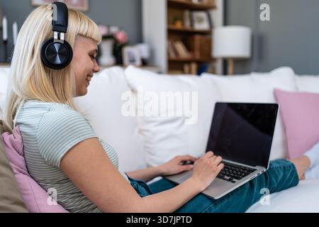 Lächelnde junge Frau mit Kopfhörern, die auf dem Sofa sitzen, während sie mit dem Laptop Musik und entspannter Bildschirmzeit beim Umschalten zwischen Online-Hengst genießt Stockfoto