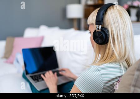 Lächelnde junge Frau mit Kopfhörern, die auf dem Sofa sitzen, während sie mit dem Laptop Musik und entspannter Bildschirmzeit beim Umschalten zwischen Online-Hengst genießt Stockfoto