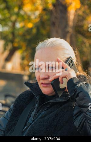 Jerewan Armenien 11.20.2025. Eine Frau spricht auf ihrem Handy, während sie auf einer Bank sitzt. Sie trägt eine schwarze Jacke und eine schwarze Weste Stockfoto