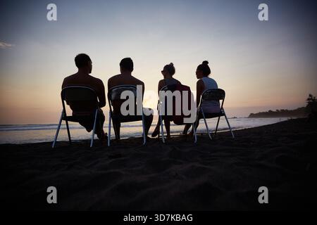 Gruppe junger Freunde, die auf Stühlen mit Blick auf das Meer sitzen und gemeinsam den Sonnenuntergang am Sandstrand beobachten, Silhouetten vor dem Himmel sichtbar, legere Sommerszene Stockfoto