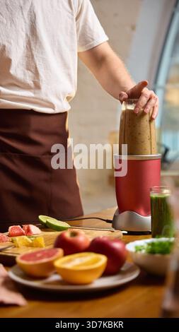Mann mischt Smoothie im kompakten Mixer mit frischem Obst und Gemüse auf dem Tisch. Stockfoto