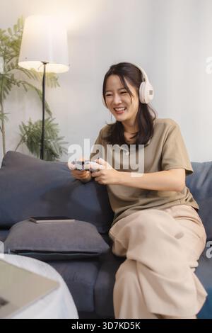 Eine junge Frau spielt zu Hause gern Videospiele, trägt Kopfhörer und hält einen Controller mit Spannung. Sie sitzt bequem auf einem Sofa und lächelt Stockfoto