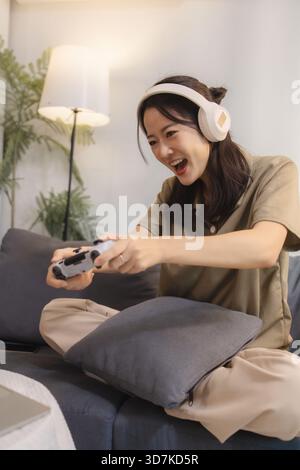 Eine junge Frau spielt zu Hause gern Videospiele, trägt Kopfhörer und hält einen Controller mit Spannung. Sie sitzt bequem auf einem Sofa und lächelt Stockfoto