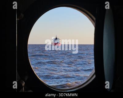 Ein großes rotes Schiff segelt auf dem Ozean, wenn man durch ein Bullauge eines anderen Schiffes sieht. Stockfoto