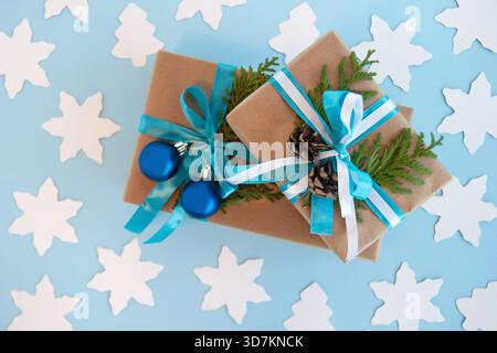 Zwei Geschenkboxen mit Bastelpapier, blauem und weißem Band und verzierten Tannenzweigen, blauen Weihnachtskugeln und Tannenzapfen auf blauem Hintergrund mit weißer Papiertanne und Stern, Ansicht von oben. Weihnachtsgeschenke. Stockfoto
