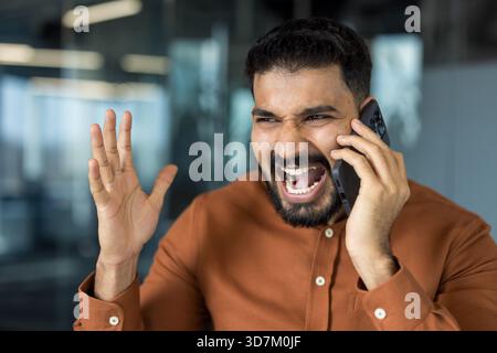 Junger, gestresster indischer Mann, der Wut und Frustration ausdrückt, während er ein intensives Telefongespräch führt und mit der Hand in einer modernen Büroumgebung Gesten macht Stockfoto