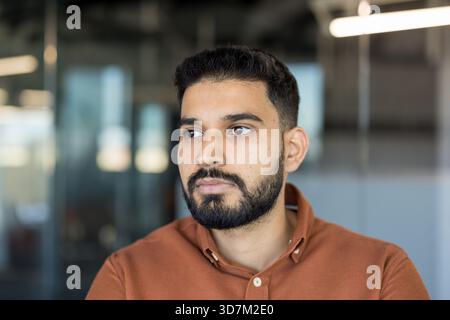 Junger Mann mit Bart und ernsthaftem Ausdruck, der tief in Gedanken zur Seite blickt und Konzepte der Planung, Vision, Kontemplation und Zukunftsorientierung in einer modernen Büroumgebung repräsentiert Stockfoto