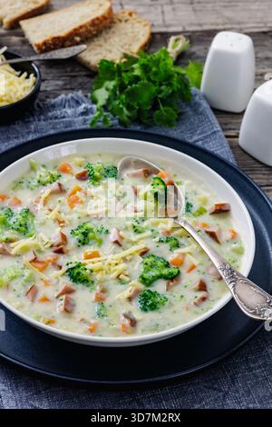 Wurstgemüse Chowder, dicke cremige Suppe mit Brokkoli, Karotten, Kartoffeln in weißer Schüssel auf rustikalem Holztisch mit Brotscheiben, vertikal Stockfoto
