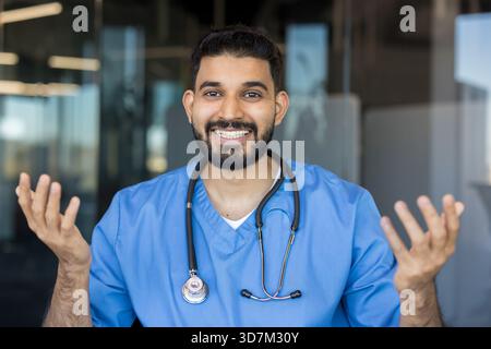 Indischer männlicher Arzt mit blauen Peelings und einem Stethoskop um den Hals, stehend mit erhobenen Händen in einer medizinischen Umgebung und lächelte selbstbewusst in die Kamera Stockfoto