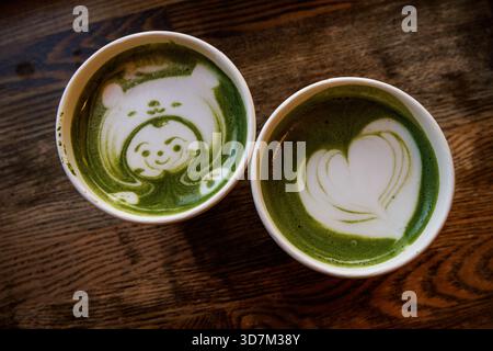 Blick von oben auf zwei grüne Matcha Lattes auf Holztisch, einer mit einem lächelnden Gesicht und einer mit Herzdesign. Stockfoto