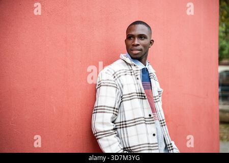 Ein junger Mann lehnt sich lässig an eine knallrote Wand und trägt eine trendige Flanelljacke mit einem farbenfrohen Pullover darunter. Sein selbstbewusster Ausdruck Stockfoto