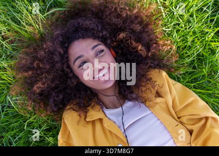 Junge Frau mit natürlichen lockigen Haaren, die auf grünem Gras liegt, lächelt, während sie Musik mit Kopfhörern hört und sich entspannt und glücklich fühlt Stockfoto