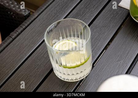 Fast leeres Glas Limettenwasser auf dunklem Tisch im Freien, klarer gerippter Tumbler mit Zitrusscheibe und schmelzendem Eis, erfrischendes Sommergetränk im Café Ter Stockfoto