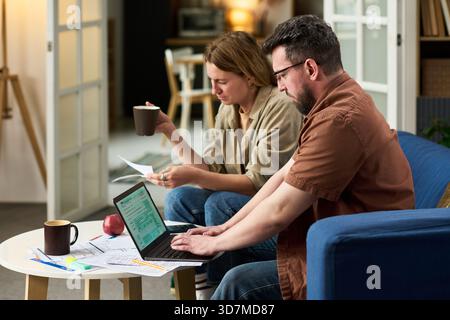 Kaukasier mittleren Alters, der Laptop benutzt, während Kaukasierin mittleren Alters Dokumente hält und Kaffee trinkt, sowohl Finanzpapiere überprüft als auch Steuern zu Hause berechnet Stockfoto