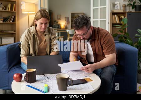 Kaukasische junge Frau und kaukasischer junger Mann sitzen auf dem Sofa, überprüfen Steuerdokumente und benutzen Laptop, konzentrieren sich auf Papierkram mit Kaffeetassen und Finanzformularen auf dem Tisch Stockfoto