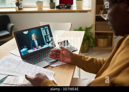 Schwarzer Mann mittleren Alters, der Steuerdokumente hält, die junge Erwachsene Kaukasierin per Videoanruf auf dem Laptop konsultieren und Finanzpapiere mit dem Rechner auf dem Tisch diskutieren Stockfoto