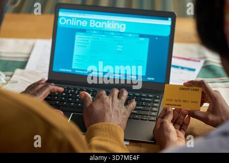Schwarzer Mann, der Laptop für Online-Banking benutzt, während er seine Kreditkarte hält, am Schreibtisch sitzt und einen weiteren unterstützenden, digitalen Steuerzahlungsprozess auf dem Computerbildschirm anzeigt Stockfoto